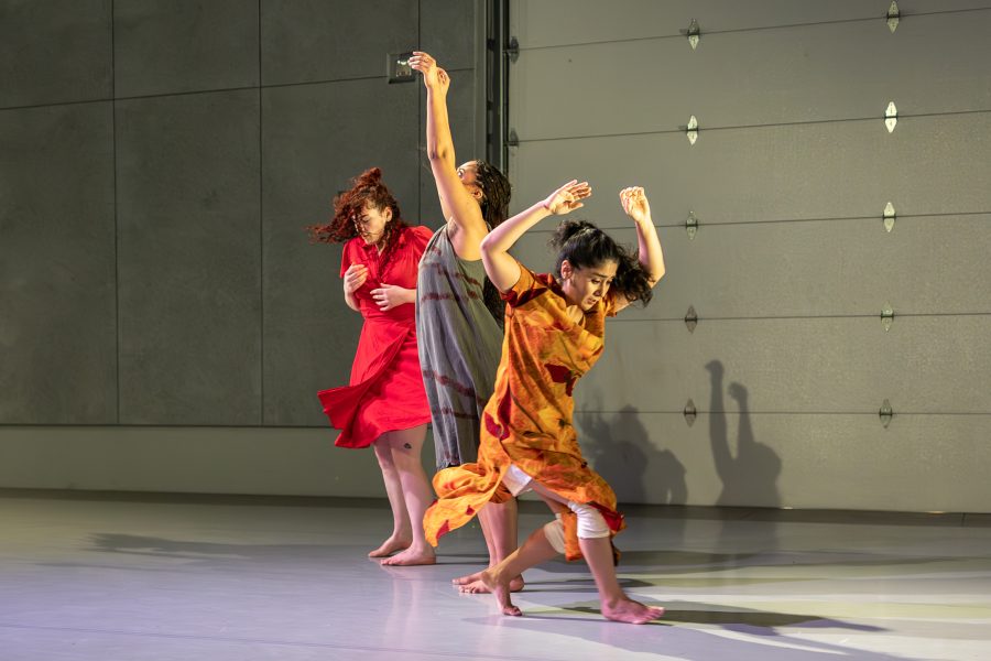 Three barefoot dancers in colorful dresses perform expressive movements on a light gray stage, with a large industrial-style wall in the background. Dramatic lighting casts shadows and highlights their poses, evoking the spirit of Braiding Futures as they weave together stories through every gesture.