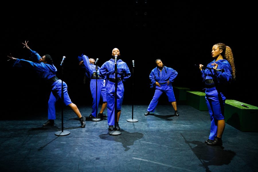 Five performers in matching blue outfits pose dynamically on stage with four microphones, each striking a unique stance. The background is dark, highlighting the performers under bright stage lighting.