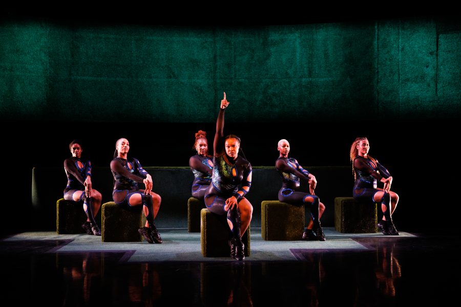 Six performers in matching black outfits sit and pose on green cubes on stage, evoking the signature style seen in many ogemdi ude major productions. One person in the center raises their hand, drawing focus under a dark background with a large green panel.
