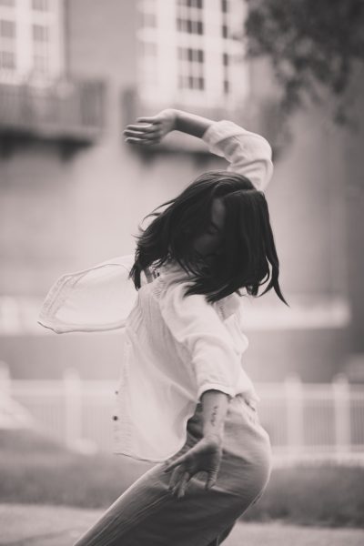 A person in a white shirt dances outdoors, arms raised and hair flowing, embodying the spirit of Braiding Futures, with a blurred building in the background. The photo is in black and white.