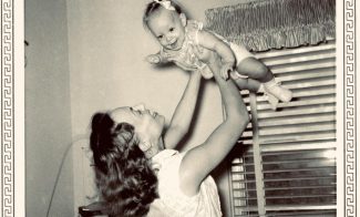 A woman embodying the spirit of motherhood, dressed in a vintage gown, lifts a smiling baby above her head in a nursery near a crib and a window with striped curtains. Reminiscent of Suzanne Bocanegra’s evocative storytelling, the photo is framed by a decorative border.