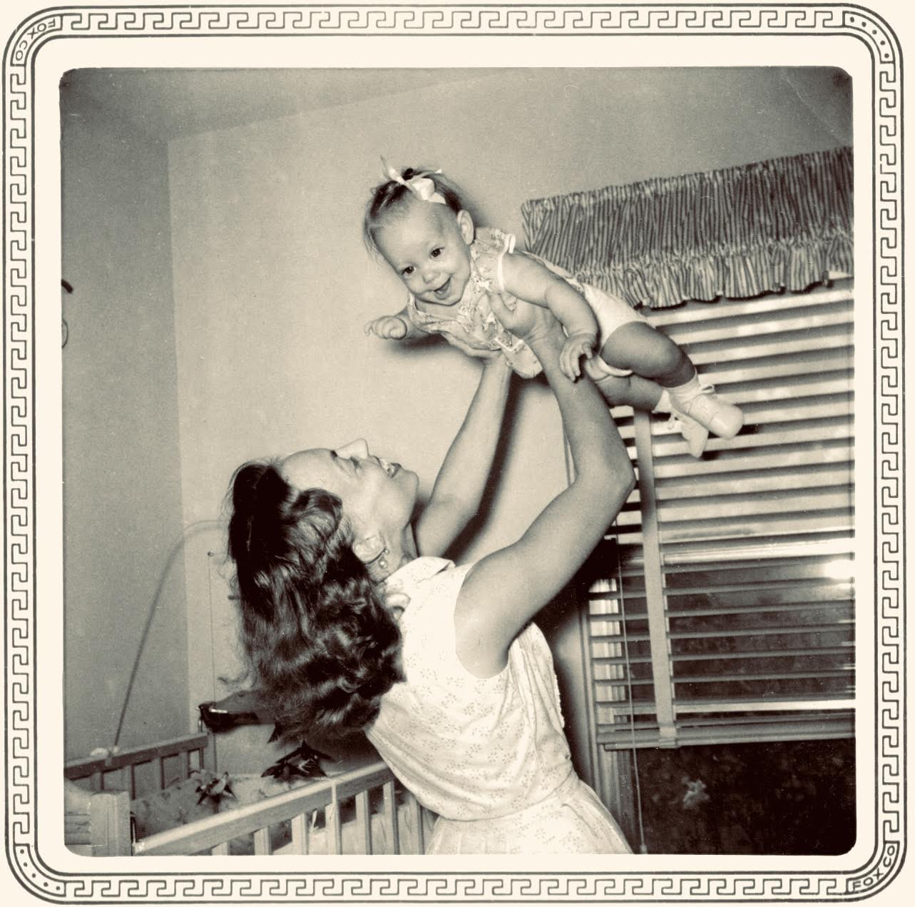 A woman embodying the spirit of motherhood, dressed in a vintage gown, lifts a smiling baby above her head in a nursery near a crib and a window with striped curtains. Reminiscent of Suzanne Bocanegra’s evocative storytelling, the photo is framed by a decorative border.