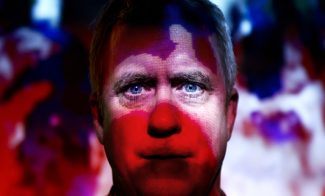 A man stands in front of a vibrant, abstract background with red and blue light patterns projected onto his face, creating a dramatic and colorful effect.