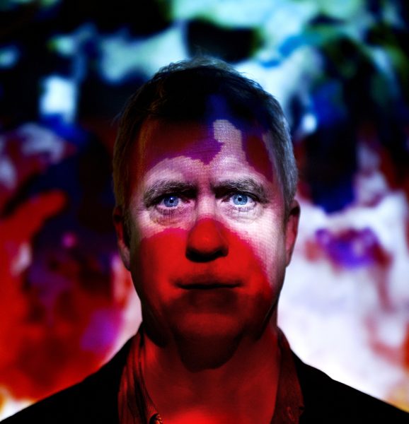 A man stands in front of a vibrant, abstract background with red and blue light patterns projected onto his face, creating a dramatic and colorful effect.
