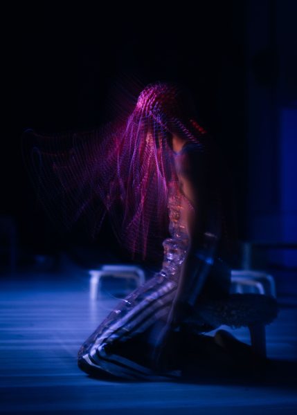 A person with glowing pink braids kneels on the floor in a dimly lit space, captured in motion blur with blue and purple lighting, creating an ethereal, surreal effect.