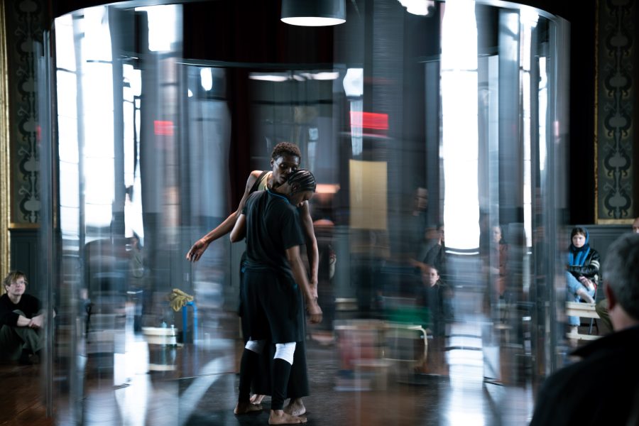 Two dancers stand close together inside a transparent cylindrical structure, with blurred motion around them. Several people sit and watch in the background in a dimly lit room with ornate walls.