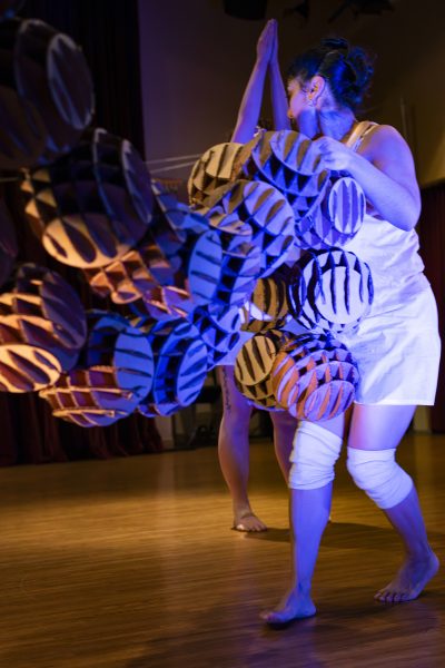 A woman in a white outfit performs a dance with large, geometric, striped props under dramatic blue and purple lighting on a wooden stage, artfully braiding futures through movement and form.