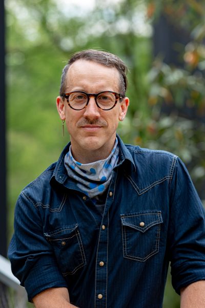 A person with short brown hair, glasses, a mustache, and an earring stands outside wearing a dark denim shirt and a patterned scarf around their neck.