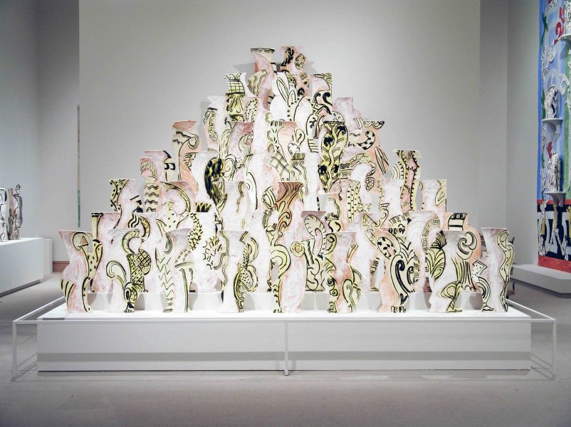 A large pyramid of painted ceramic vases with abstract black and gold designs is displayed on a white pedestal in an art gallery with neutral walls.