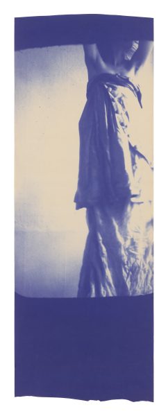 A blue-toned photo of a person standing against a wall, shown from shoulders down, wearing a long, textured skirt or dress. Their arms are crossed over their chest. The image has a soft and artistic feel.