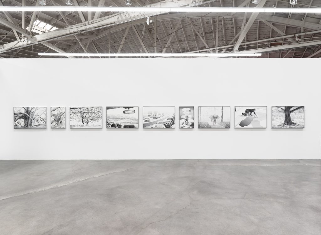A row of black-and-white drawings hangs on a white gallery wall in a spacious room with exposed beams and a polished concrete floor. The artworks depict various scenes, mostly involving trees and figures.