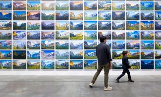 An adult and a child walk past an ICA events gallery wall covered in a grid of landscape paintings depicting mountains and valleys.