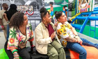 Three people sit together on a colorful bench at an indoor art exhibit during one of the vibrant ICA events, engaging and smiling. Bright artwork and more visitors are visible in the background.