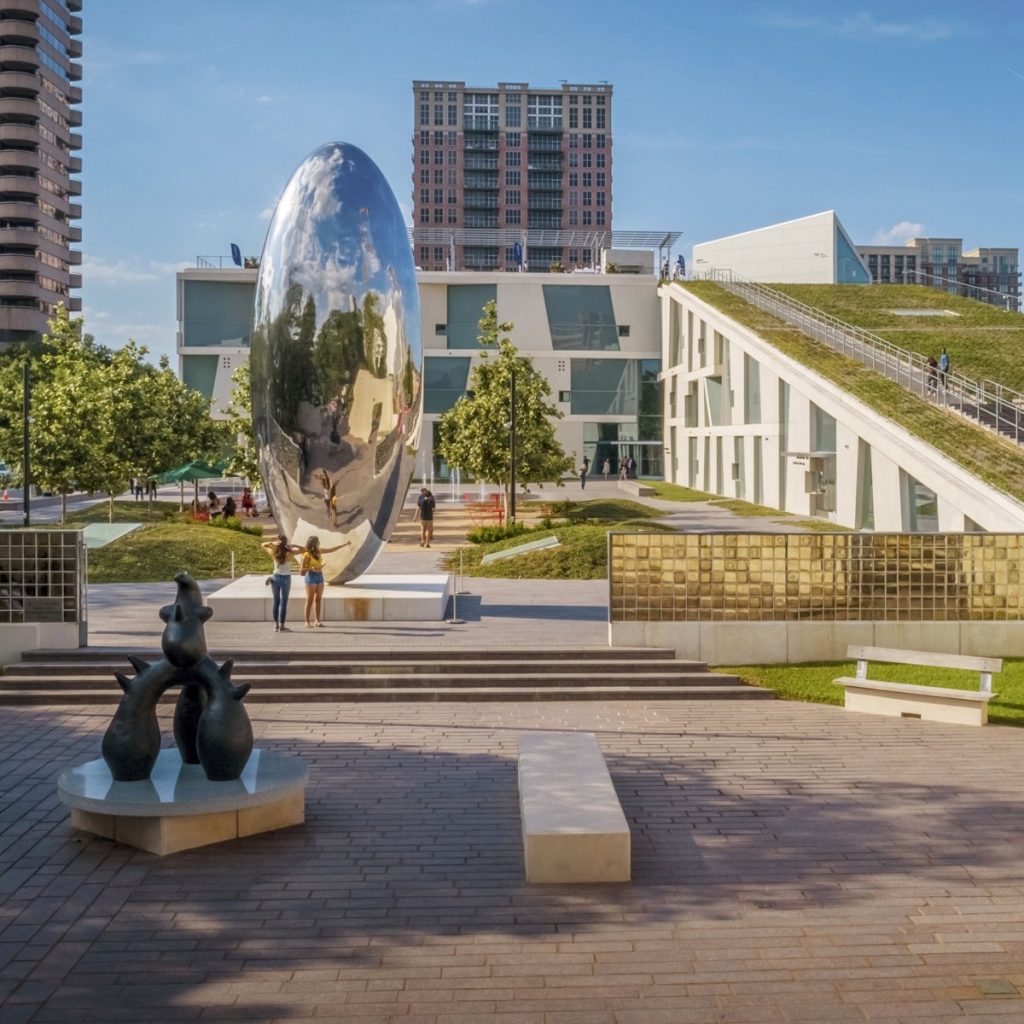 A large, shiny, bean-shaped sculpture stands in a plaza surrounded by modern buildings, trees, and people—much like the striking installations often seen at ICA Houston. In the foreground are abstract animal statues and benches. The sky is clear and blue.