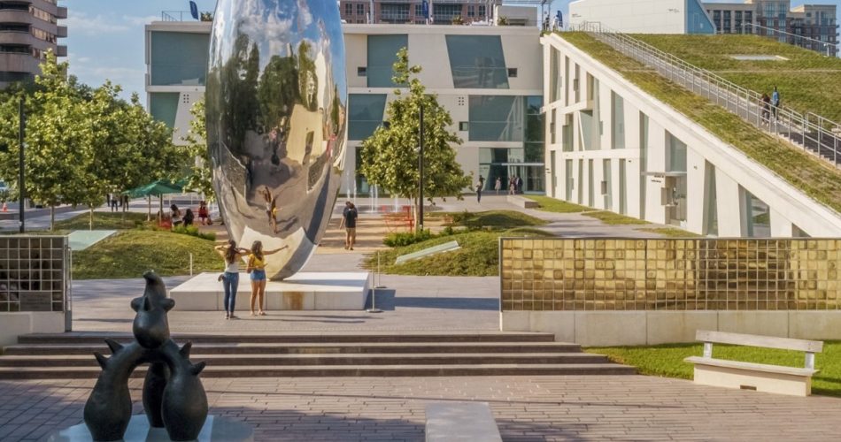 A large, shiny, bean-shaped sculpture stands in a plaza surrounded by modern buildings, trees, and people—much like the striking installations often seen at ICA Houston. In the foreground are abstract animal statues and benches. The sky is clear and blue.