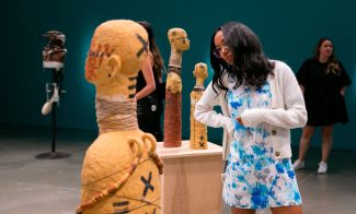 A woman in a white cardigan and blue floral dress observes a sculpted figure in an art gallery, perhaps drawing inspiration for an upcoming Art21 Educator Workshop. Other sculptures and visitors are seen in the background against teal walls.