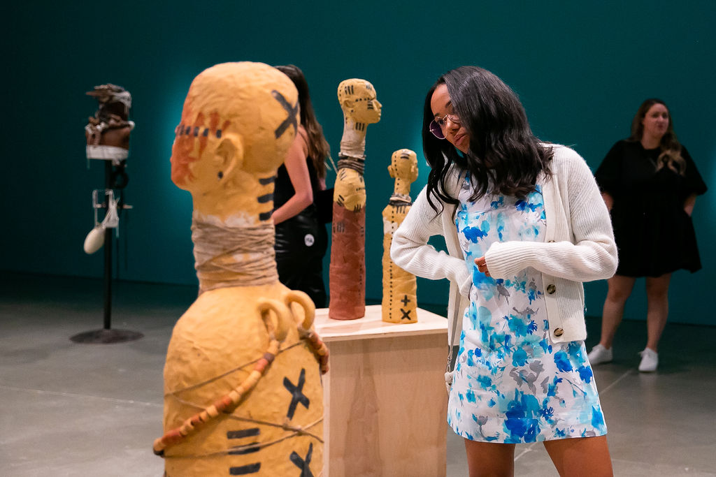 A woman in a white cardigan and blue floral dress observes a sculpted figure in an art gallery, perhaps drawing inspiration for an upcoming Art21 Educator Workshop. Other sculptures and visitors are seen in the background against teal walls.