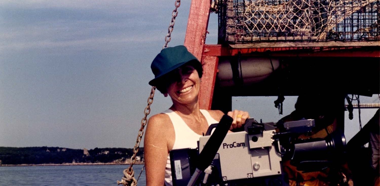 A person in a teal cap and sleeveless top smiles while holding a ProCam video camera on a boat, with lobster traps and calm water in the background—capturing moments reminiscent of Joan Logue’s iconic video portraits featured at ICA Boston.