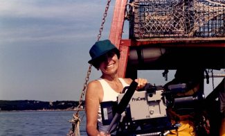 A person in a teal cap and sleeveless top smiles while holding a ProCam video camera on a boat, with lobster traps and calm water in the background—capturing moments reminiscent of Joan Logue’s iconic video portraits featured at ICA Boston.