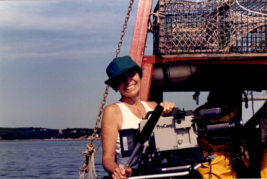 A person in a teal cap and sleeveless top smiles while holding a ProCam video camera on a boat, with lobster traps and calm water in the background—capturing moments reminiscent of Joan Logue’s iconic video portraits featured at ICA Boston.