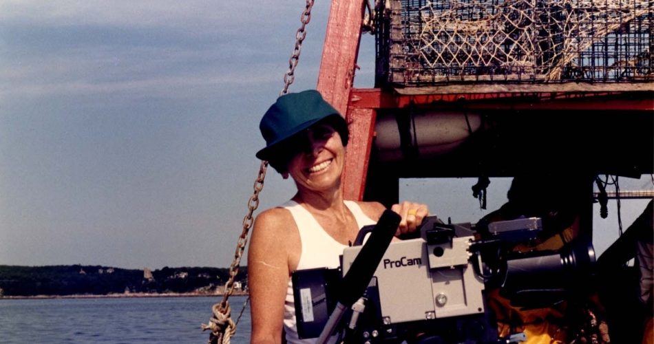A person in a teal cap and sleeveless top smiles while holding a ProCam video camera on a boat, with lobster traps and calm water in the background—capturing moments reminiscent of Joan Logue’s iconic video portraits featured at ICA Boston.