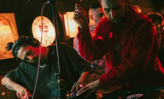 Two DJs perform in a dimly lit club during First Fridays at ICA Boston, one mixing vinyl records while the other adjusts equipment. Warm lights and a focused crowd create an energetic nighttime atmosphere.