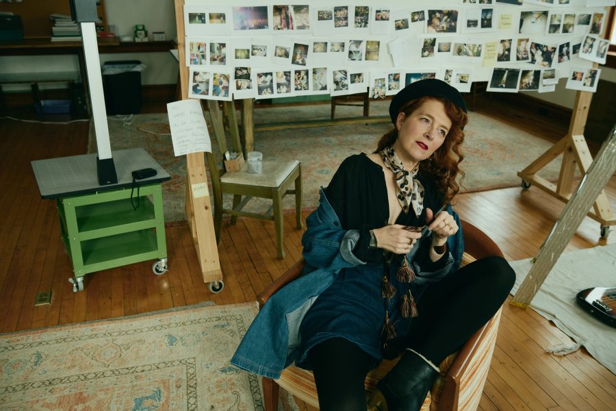 A woman with wavy red hair sits in a chair, wearing a black hat and scarf, in an art studio with photos and notes pinned to boards behind her on wooden stands. The room has wooden floors and a patterned rug, giving the impression that she might be preparing her thoughts for an upcoming ICA book talk.