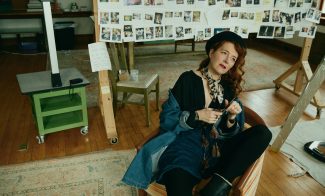 A woman with long red hair sits in a chair in an art studio, wearing a hat and scarf. Behind her, dozens of photos are clipped to boards, and the floor has a patterned rug. The room feels creative and relaxed—like the perfect setting for an ICA book talk.