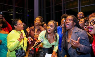 A group of people sing passionately together at an indoor event, likely part of the lively First Fridays at ICA Boston; one woman holds a microphone and others appear excited and joyful, with colorful outfits and expressive faces.