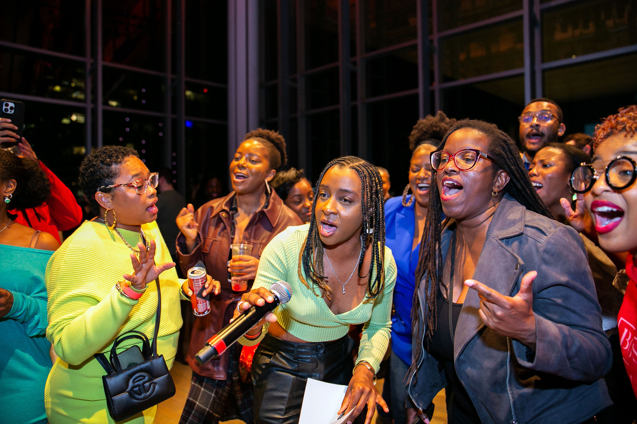 A group of people sing passionately together at an indoor event, likely part of the lively First Fridays at ICA Boston; one woman holds a microphone and others appear excited and joyful, with colorful outfits and expressive faces.