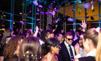 A crowd of people in formal attire dance and celebrate under purple lighting, with flowers falling from above in a large glass-walled venue at night.