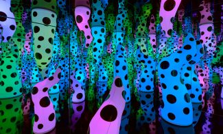 Colorful, polka-dotted sculptural forms in pink, green, and blue fill a mirrored room, creating endless reflections and a sense of infinite space and repetition.