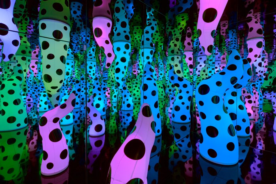Colorful, polka-dotted sculptural forms in pink, green, and blue fill a mirrored room, creating endless reflections and a sense of infinite space and repetition.