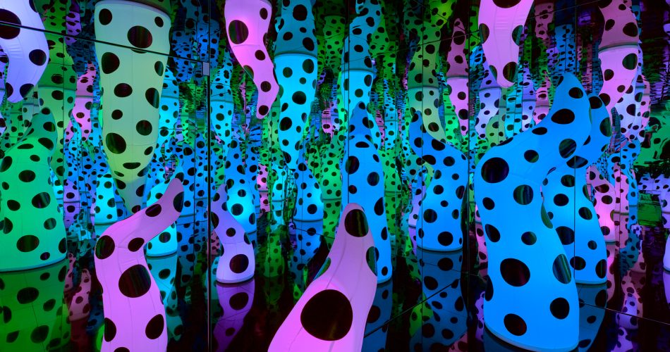 Colorful, polka-dotted sculptural forms in pink, green, and blue fill a mirrored room, creating endless reflections and a sense of infinite space and repetition.