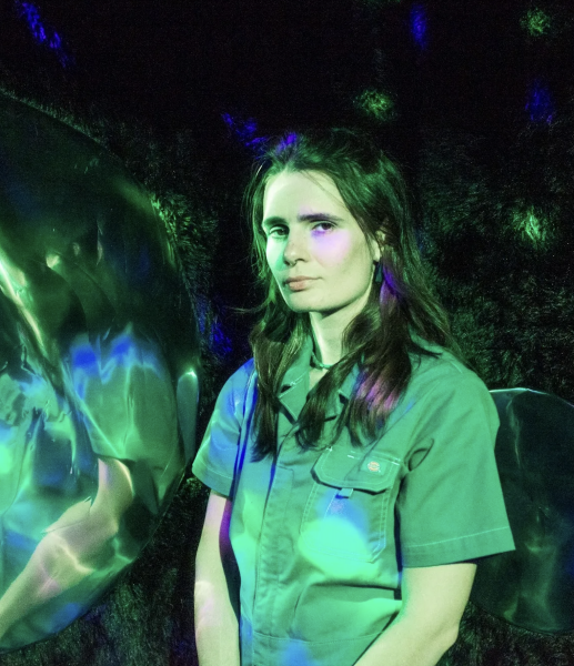 A person wearing a green short-sleeved jumpsuit stands next to a large, reflective metallic sphere under green and blue lighting at the ICA Halloween party.