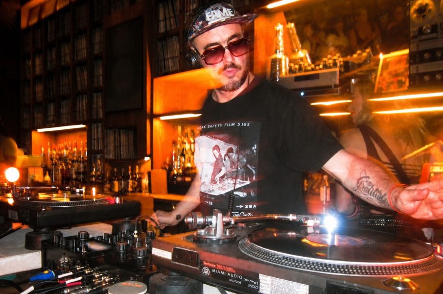 A DJ wearing sunglasses and a black t-shirt is mixing vinyl records in a dimly lit bar with a warm, orange glow—capturing the electrifying vibe of an ICA Halloween Party. Shelves filled with records are visible in the background.