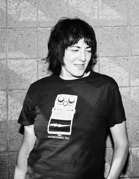 Black and white photo of a person with short dark hair, wearing a t-shirt featuring a guitar pedal graphic and the words Guitar Bar, Hoboken, NJ, standing in front of a textured brick wall.