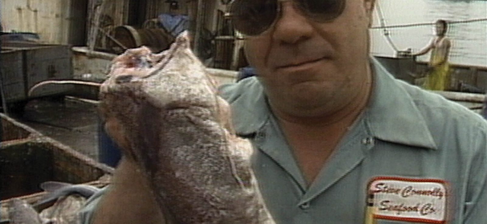 A man in sunglasses holds a large fish on a dock, wearing a blue shirt with a name patch that reads Steve Connolly Seafood Co. Boats and seafood are visible in the background.