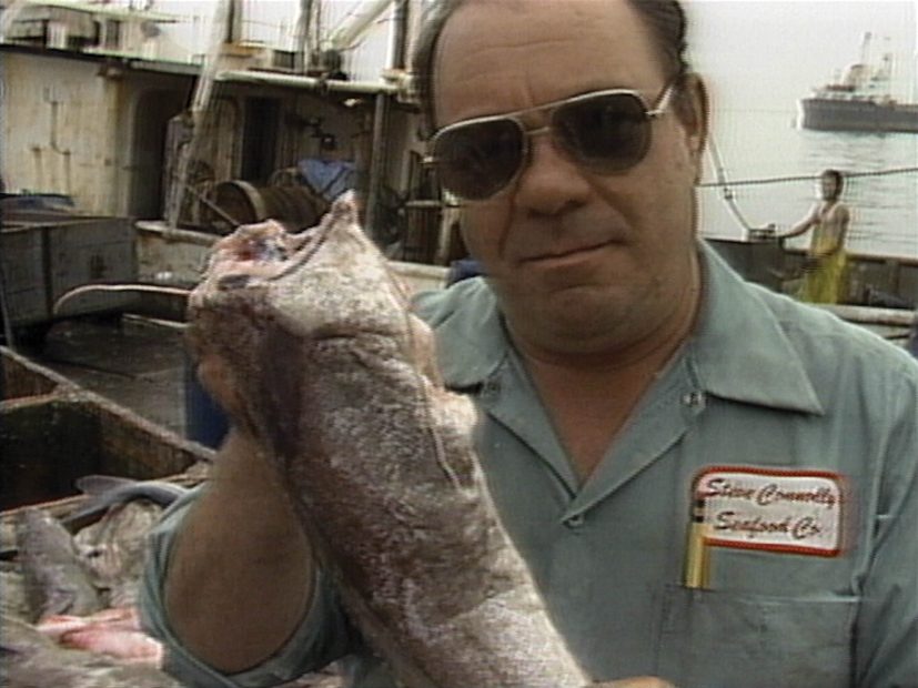 A man in sunglasses holds a large fish on a dock, wearing a blue shirt with a name patch that reads Steve Connolly Seafood Co. Boats and seafood are visible in the background.
