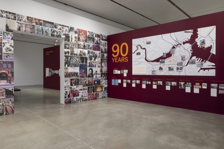 Museum exhibition gallery featuring a wall covered in photo collages, a 90 Years title, a large map of Boston with historic site markers, and smaller framed photographs with descriptive captions.