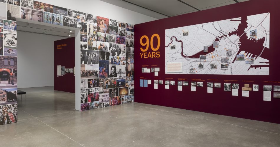 Museum exhibition gallery featuring a wall covered in photo collages, a 90 Years title, a large map of Boston with historic site markers, and smaller framed photographs with descriptive captions.