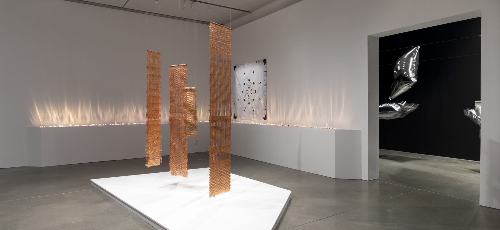 Three metallic, textured rectangular panels hang from the ceiling above a white platform in a gallery, with a line of glowing lights along the wall behind them.