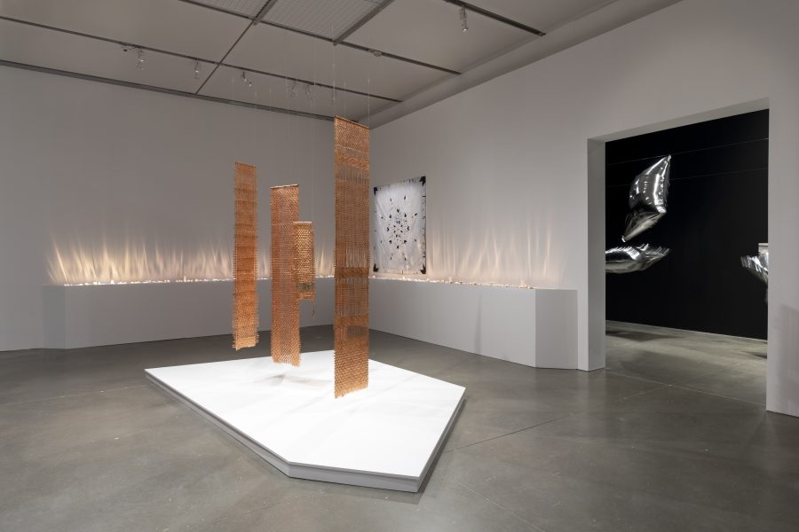 Three metallic, textured rectangular panels hang from the ceiling above a white platform in a gallery, with a line of glowing lights along the wall behind them.