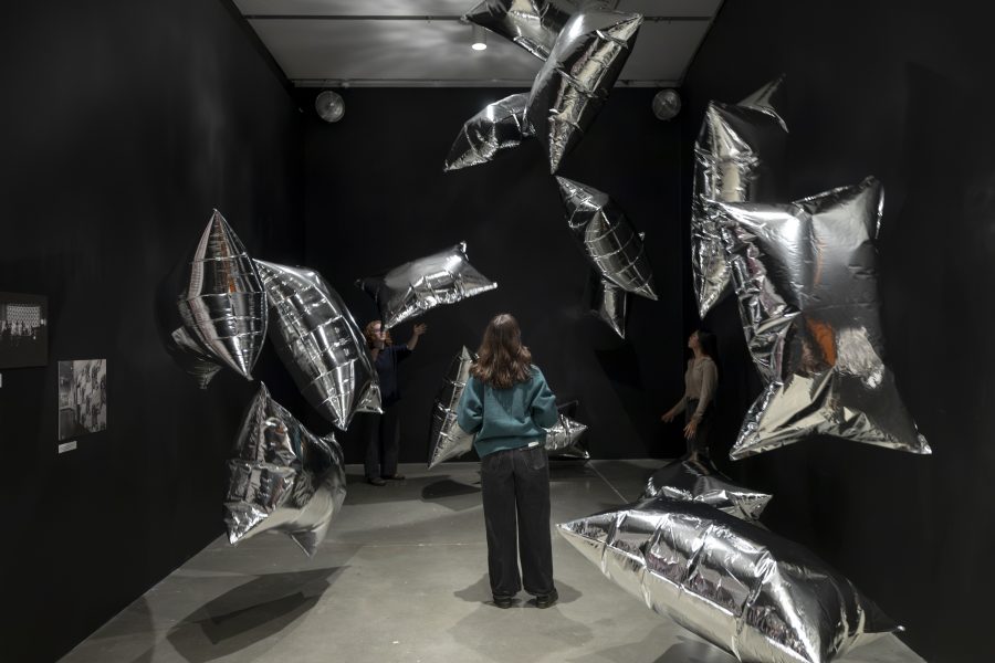 Three people in a black room surrounded by large, floating silver pillow-shaped balloons.