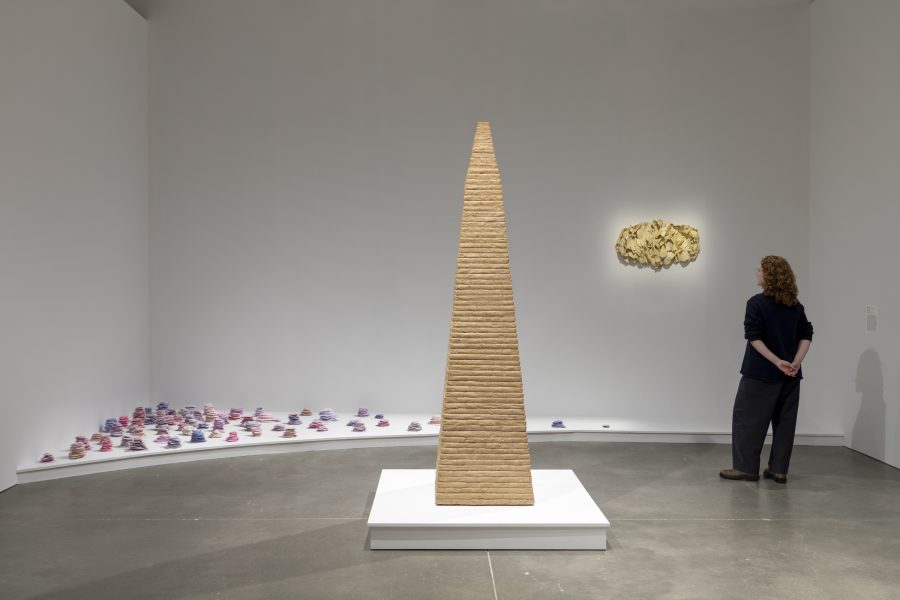 A person stands in a gallery looking at a tall, tiered, triangular sculpture, with small colorful shapes arranged on the floor and a textured wall piece nearby.