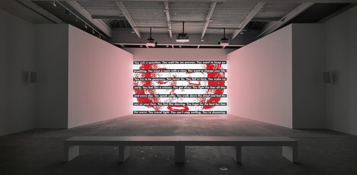 A modern art gallery with a bench facing a large wall display, reminiscent of Barbara Kruger, featuring text covered in red paint like an abstract American flag at ICA Boston under bright industrial lighting.