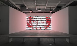 A modern art gallery with a bench facing a large wall display, reminiscent of Barbara Kruger, featuring text covered in red paint like an abstract American flag at ICA Boston under bright industrial lighting.