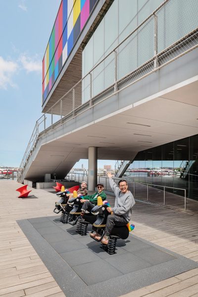 Three people sit on animal-shaped spring riders in front of a modern building with a colorful upper facade, wooden deck, and clear sky in the background.