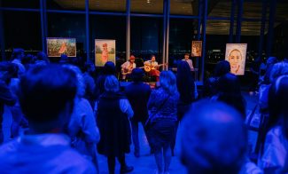 A musician plays guitar and sings on a small stage in front of an audience in a dimly lit room with art pieces displayed on stands.