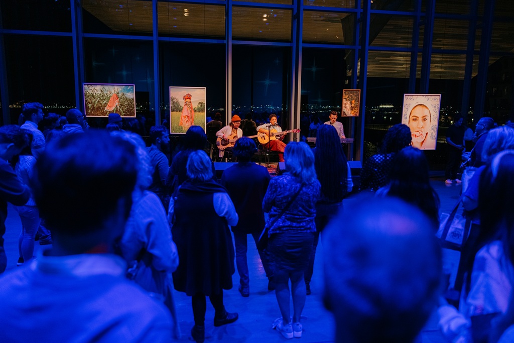 A musician plays guitar and sings on a small stage in front of an audience in a dimly lit room with art pieces displayed on stands.
